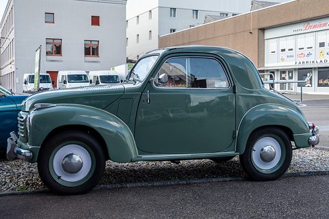 Grey-green small saloon side-on on gravel in front of an industrial building. Opens a larger view