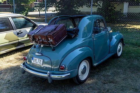 Grey small car from the left rear on grass, a suitcase on the tail rack and a fence behind. Opens a larger view