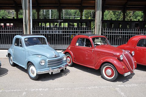 Light blue small saloon and a red small saloon with a tall grille parked side by side under a shelter. Opens a larger view