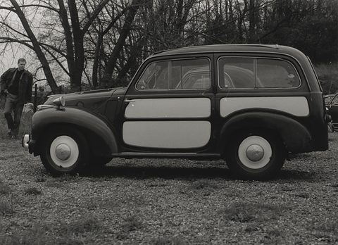 Black and white side view of a dark small estate with light side panels parked on grass under bare trees. Opens a larger view