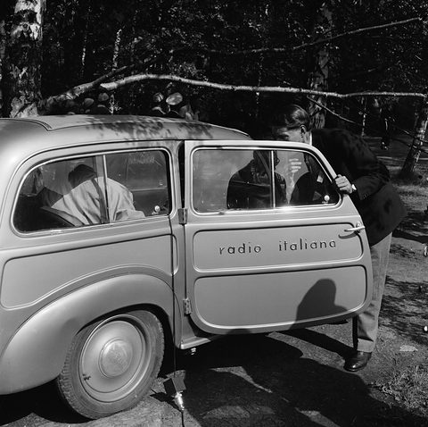 Black and white: a small estate lettered radio italiana on its door, a man leaning in, trees behind.