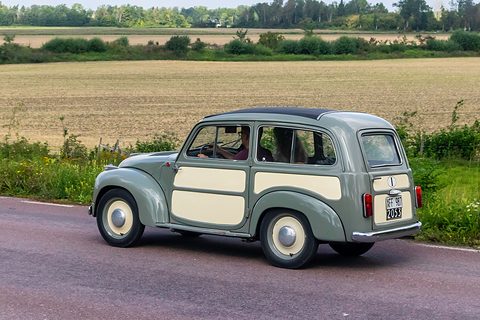 Grey small estate with cream panels driving along a road beside a field. Opens a larger view