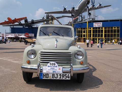 Front of a pale green small estate with rally plate 109, aircraft displayed on the museum roof behind. Opens a larger view