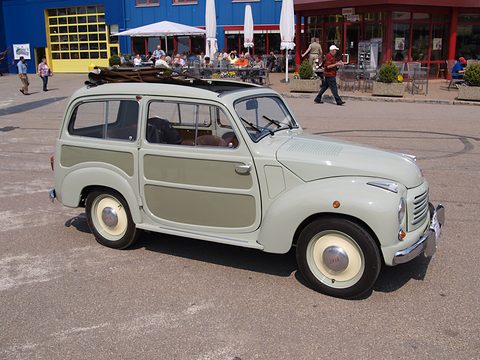 Pale green small estate side-on with a roof rack, on a square before a museum building. Opens a larger view