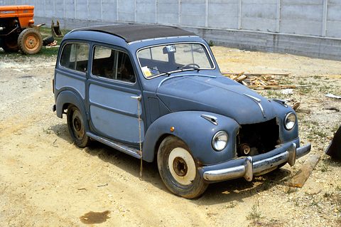 Faded blue small estate without a grille parked on sandy ground in front of a fence. Opens a larger view