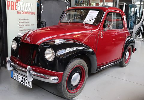 Red small saloon with black wings and red wheels in a showroom, a dealer’s banner at the left. Opens a larger view