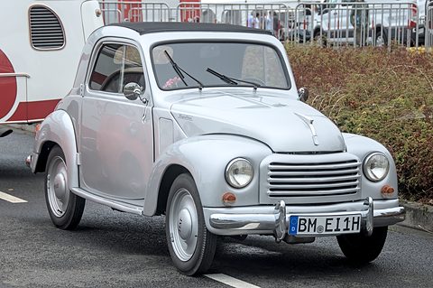 Silver small saloon with a horizontal-bar grille parked on tarmac beside a hedge. Opens a larger view
