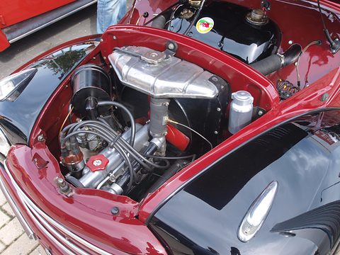 Engine bay under a raised maroon bonnet: aluminium cylinder head, carburettor and ignition leads. Opens a larger view