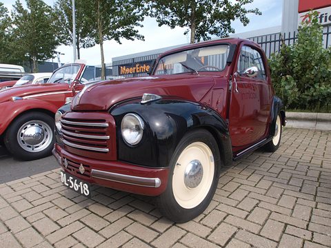 Maroon small saloon with black wings, faired-in headlamps and a horizontal-bar grille, on brick paving. Opens a larger view