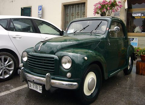 Dark green small saloon parked beside a white modern car in a village street with flowers. Opens a larger view