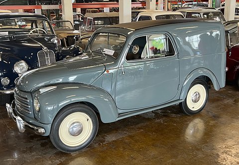 Grey small van with blank side panels and cream wheels on a wet-looking museum floor. Opens a larger view