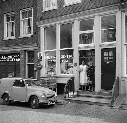 Black and white: a small van parked outside a shop where two women stand in the doorway. Opens a larger view
