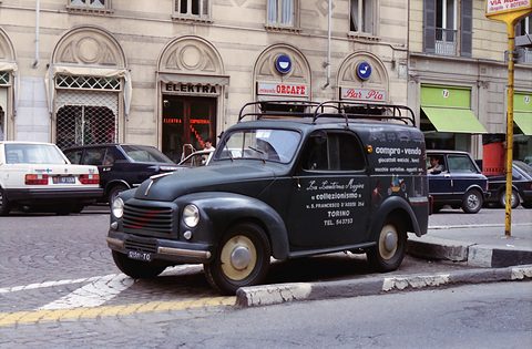 Black small van with a roof rack and white lettering, parked at the kerb before an arcaded building. Opens a larger view
