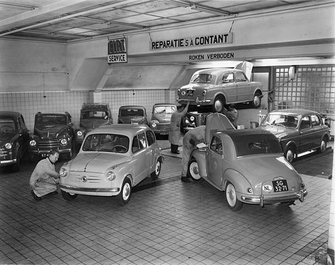 Black and white: a tiled workshop with small Fiats, one on a lift, and mechanics at work.