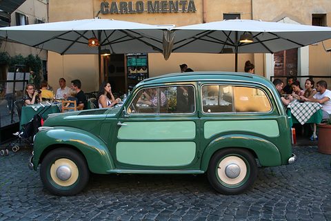Green and pale green small estate parked on cobbles in front of a restaurant terrace with umbrellas. Opens a larger view