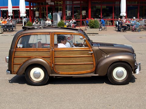 Brown small estate with varnished wooden side panels side-on in front of a museum building and café. Opens a larger view