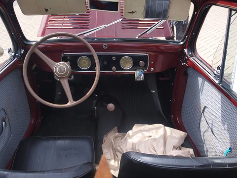Maroon dashboard with three dials, a thin steering wheel and black seats, a cloth on the floor. Opens a larger view