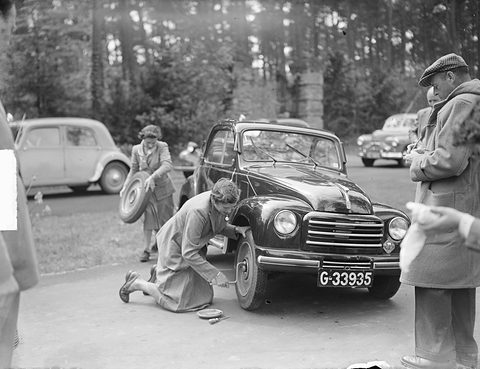 Black and white: women kneeling to change the wheel of a small saloon on a road, onlookers around. Opens a larger view