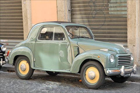 Faded pale green small saloon with yellow wheels parked on cobbles in front of a closed metal shutter. Opens a larger view