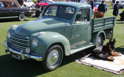 Green small pick-up on a lawn with a basket in its bed and a dog lying on a rug beside it. Opens a larger view