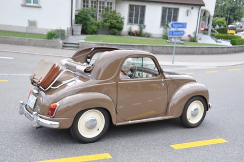 Brown small saloon from the left rear on a road, a suitcase on a rack at the tail. Opens a larger view