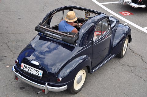 Navy small car seen from above with its roof rolled back and a driver in a straw hat at the wheel. Opens a larger view