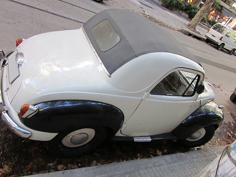 Cream small car with black wings and a dark fabric roof, seen at a tilted angle from the rear. Opens a larger view