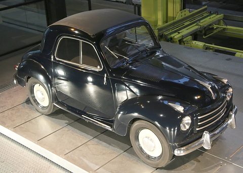 Black small saloon with a brown fabric roof on a tiled display floor, seen from above. Opens a larger view