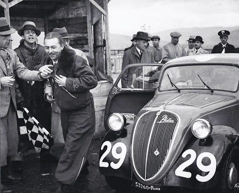 Black and white: a man in a leather jacket stepping from a small saloon numbered 29, officials and a chequered flag around him.