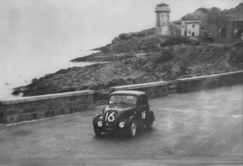 Black and white: a small saloon numbered 16 rounding a bend on a coast road below a stone tower. Opens a larger view