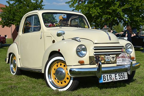 Cream small saloon with yellow wheels, whitewall tyres and rally plaques on the bumper, on grass. Opens a larger view