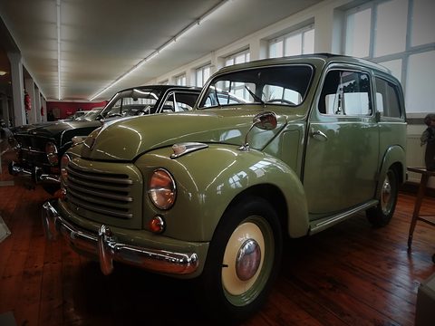 Olive green small estate with cream wheels in a long museum hall with a wooden floor. Opens a larger view