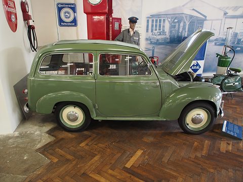 Green small estate side-on on a parquet floor, a petrol-station display and mannequin behind. Opens a larger view