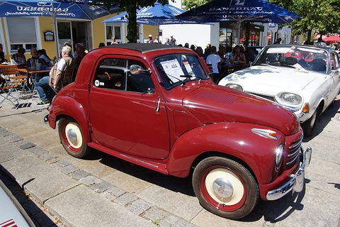 Red small saloon with cream wheels on a street, café parasols and visitors behind. Opens a larger view