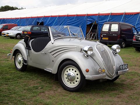 Silver open two-seater with swept chrome flashes on its wings and a tall grille, on grass. Opens a larger view