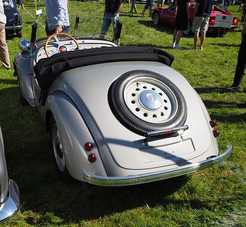 Cream open two-seater from behind with a spare wheel on its tail and a wood-rimmed wheel, on grass. Opens a larger view
