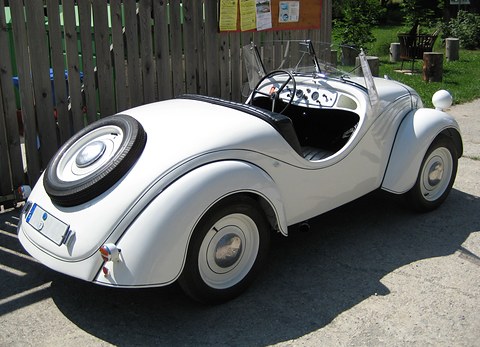 White open two-seater from the left rear with a spare wheel set into its tail. Opens a larger view