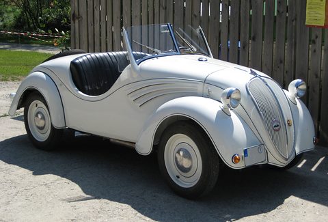 White open two-seater with swept wing flashes and black seats, beside a wooden fence. Opens a larger view