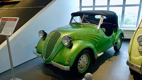 Bright green open two-seater with a black hood up and a tall grille in a white museum hall. Opens a larger view