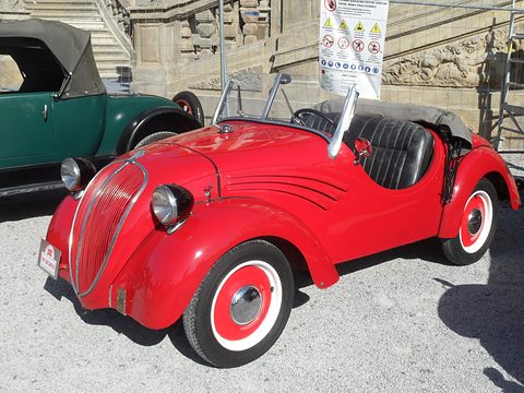 Red open two-seater with black seats and swept wing flashes on gravel, a green car behind. Opens a larger view