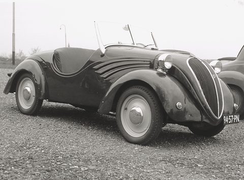 Black and white view of a dark open two-seater with swept wing flashes parked on gravel. Opens a larger view