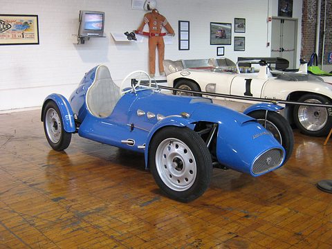 Blue open-wheeled single-seat racing car on a brick floor, a race suit hanging on the wall behind. Opens a larger view