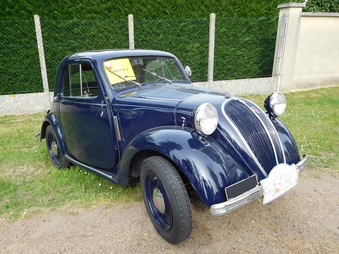 Navy small saloon with black wings, a tall grille and a card on the windscreen, before a hedge. Opens a larger view
