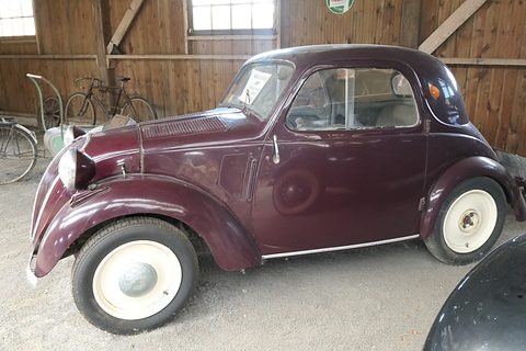 Maroon small saloon with cream wheels in a timber barn with old bicycles behind. Opens a larger view