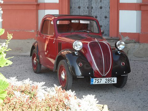 Red and black small saloon on gravel before a red and white palace, flowers in front. Opens a larger view