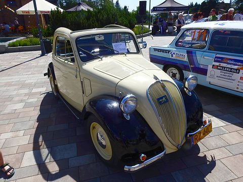 Cream small saloon with navy wings and a tall grille on paving, a rally car beside it. Opens a larger view