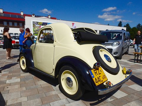 Cream small car with navy wings from the left rear, roof rolled back, on paving under a blue sky. Opens a larger view