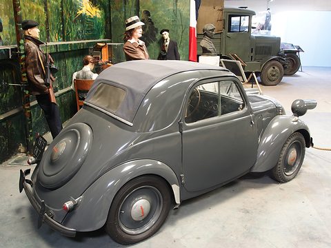 Grey small car from the left rear with its fabric roof closed, mannequins and an army truck behind. Opens a larger view