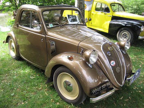 Bronze-brown small saloon with cream wheels and a badge-covered grille, parked on grass. Opens a larger view