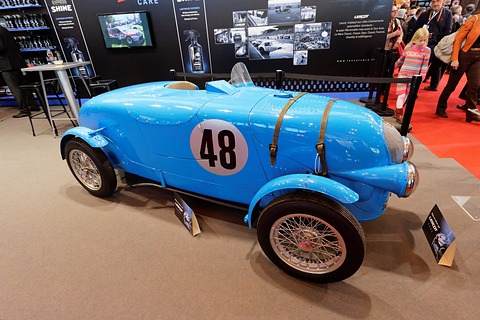 Light blue open racing car with leather bonnet straps and wire wheels on a show stand, visitors at right. Opens a larger view